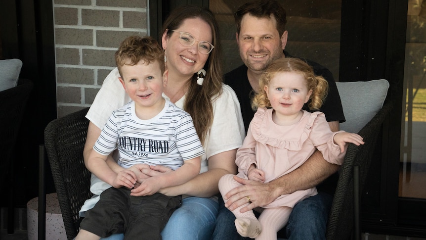 The Tucker family sit on an outdoor cane chair. Flynn sits on his mother's lap and Zoe sits on her father's lap.