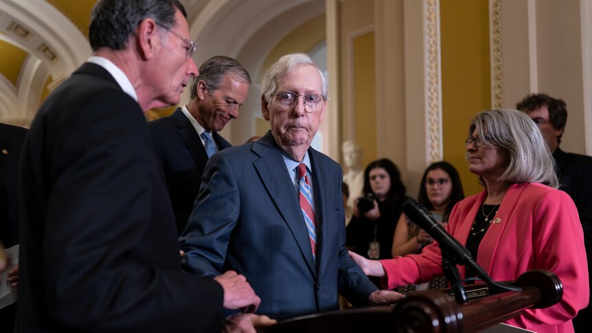 An elderly man at a podium looks bewildered while two other senators touch his arm 