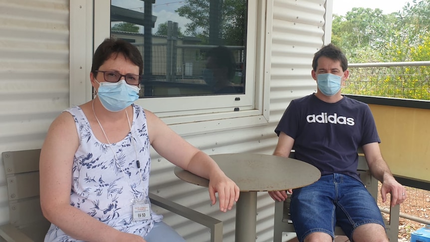 Yolandé and Hugo wearing blue surgical masks, sitting at a small table outside a basic worker's cottage.