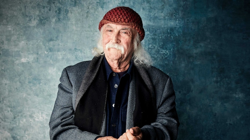 A man with grey wispy hair and moustache, wearing a red knitted beanie poses for the camera.