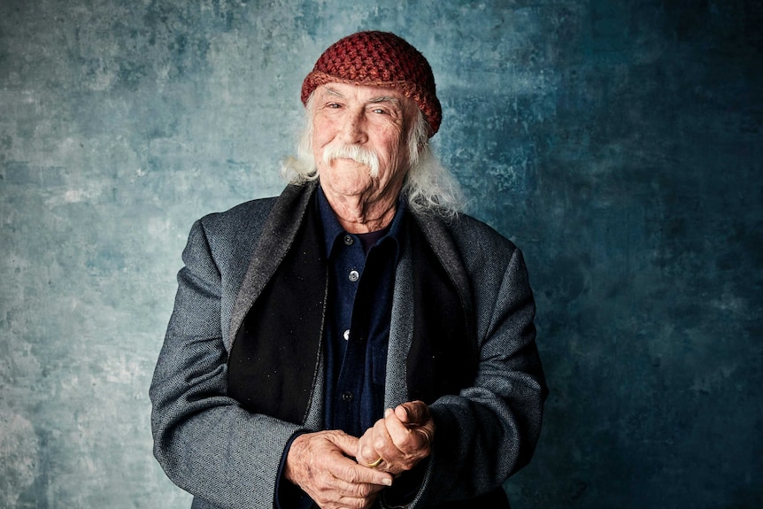 A man with grey wispy hair and moustache, wearing a red knitted beanie poses for the camera.