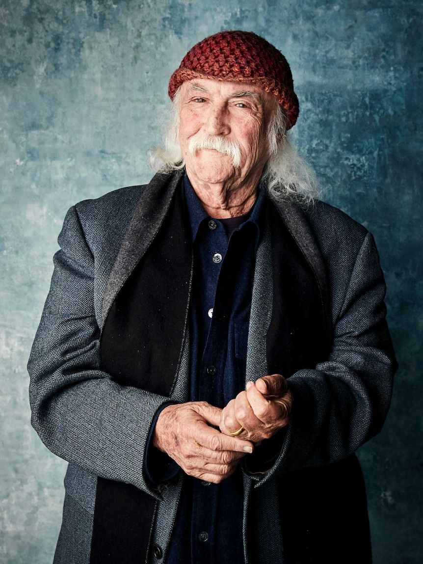 A man with grey wispy hair and moustache, wearing a red knitted beanie poses for the camera.