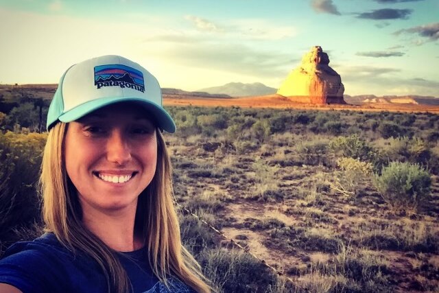 A woman smiles, with blonde hair and a blue cap. There's a humungous rock in the background with green bushes
