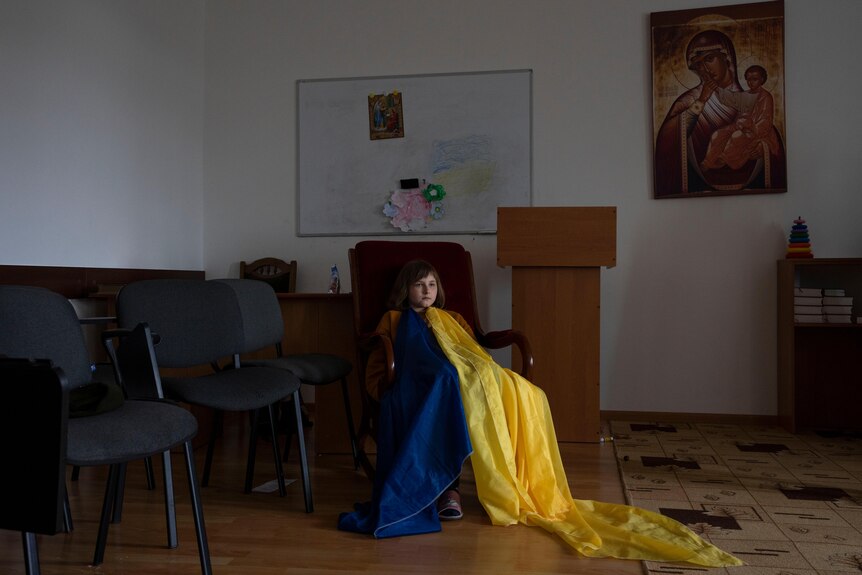 A child sits in a chair in a dimly lit room, near empty chairs. They are covered by large pieces of blue and yellow fabric.