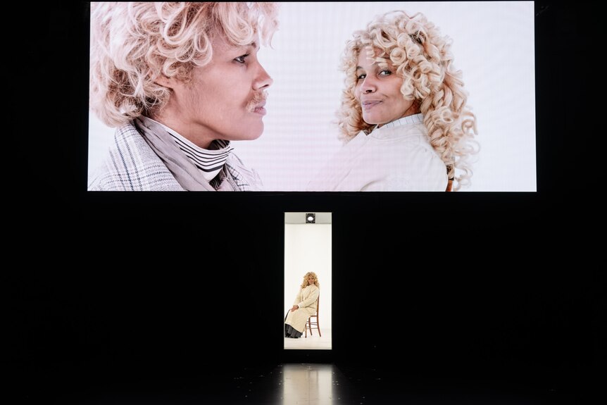 On stage, Zahra Newman, in a blonde wig, sits in a white room. Two of her are close-up on screen, one wearing a blond man's wig.