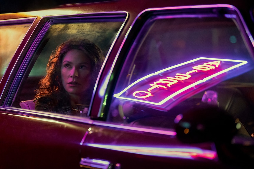 A middle-aged woman with curly hair sits in the back of a car on a neon-lit street.