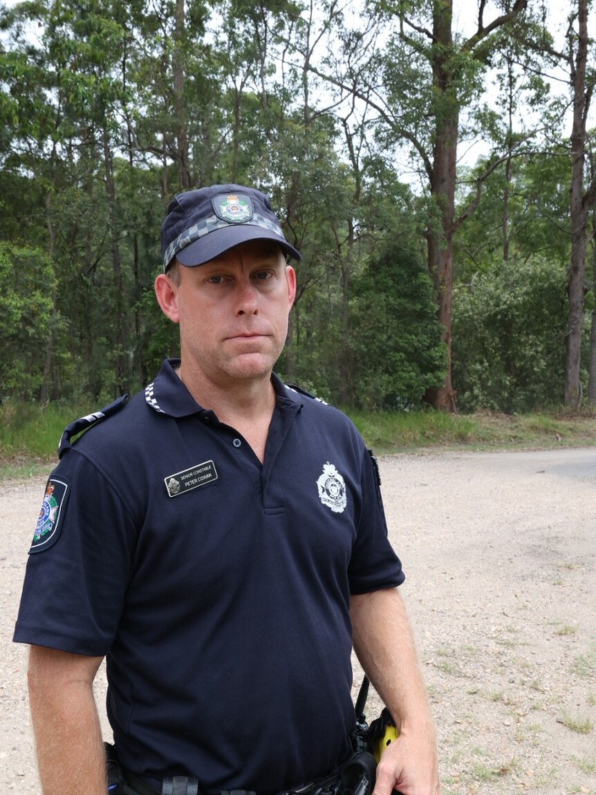 A police officer looking sombre in front of trees and a rural dirt road.