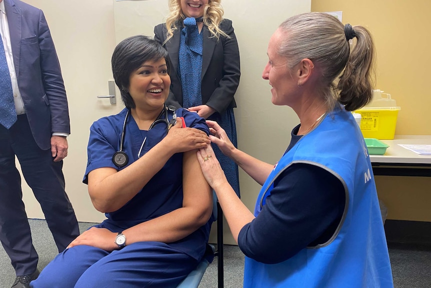 A seated woman in a nurse's uniform and a woman in a blue vest, each pressing a hand against the nurse's shoulder.