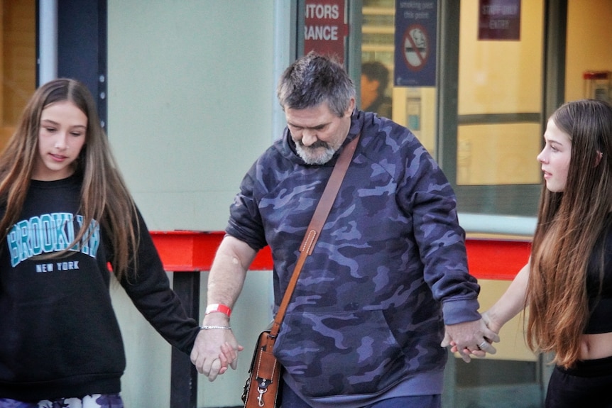 An old man walks out of hospital with a girl holding each of his hands. 