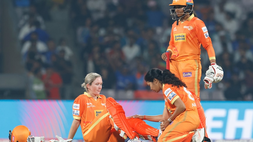 An orange-clad cricketer looks down at her knee as she sits on the ground while a medic checks out her injury.