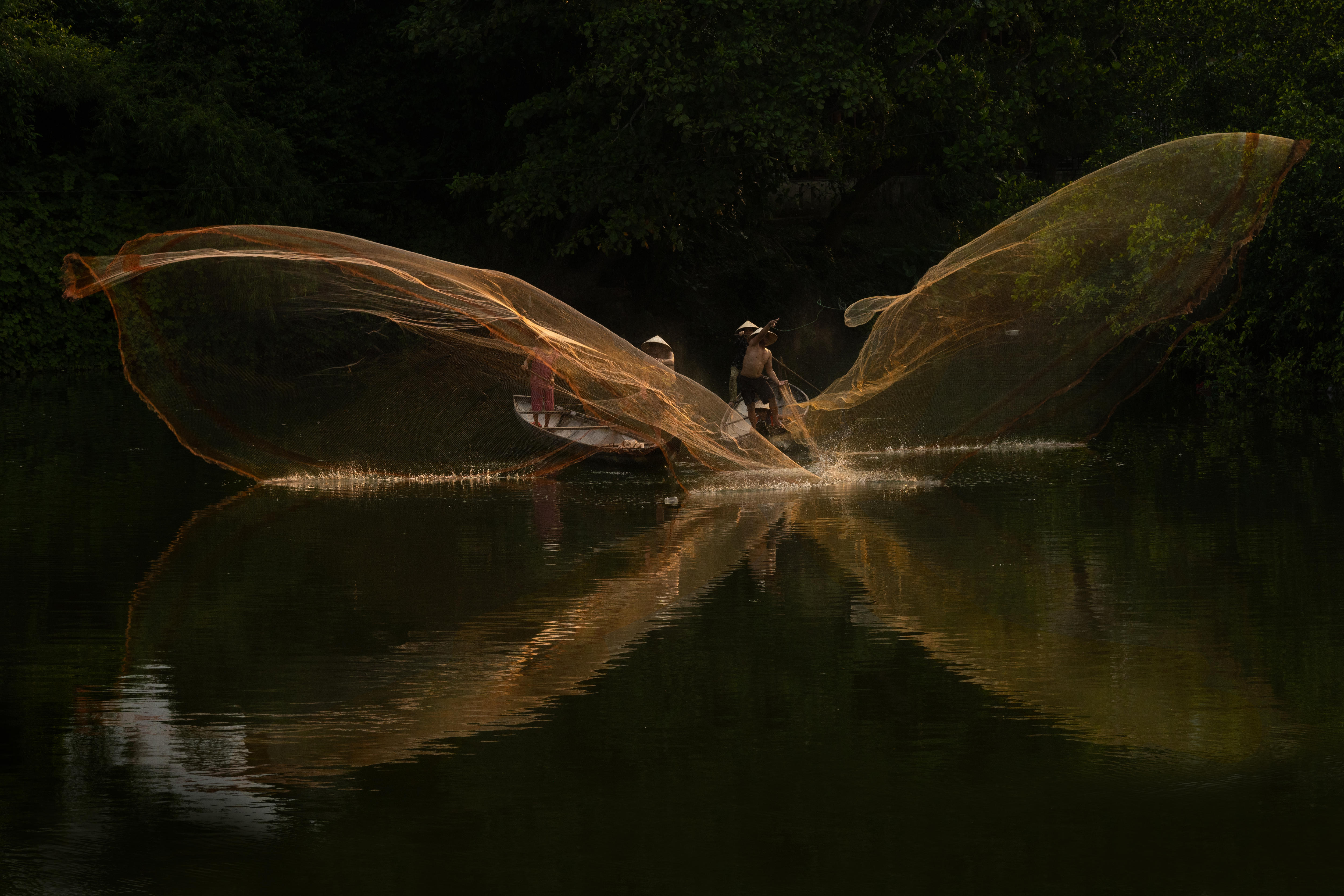 Local fishermen cast their nets from two sampan boats. The waters and sky are dark. 