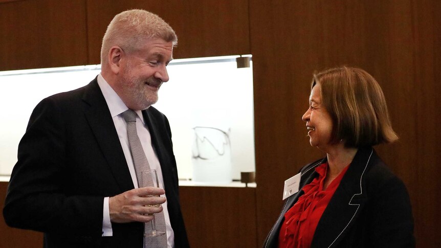 Michelle Guthrie and Mitch Fifield smile as they chat at an evening function. He is holding a champagne glass.