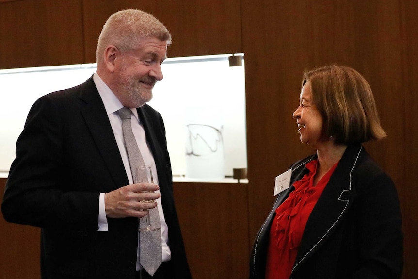 Michelle Guthrie and Mitch Fifield smile as they chat at an evening function. He is holding a champagne glass.