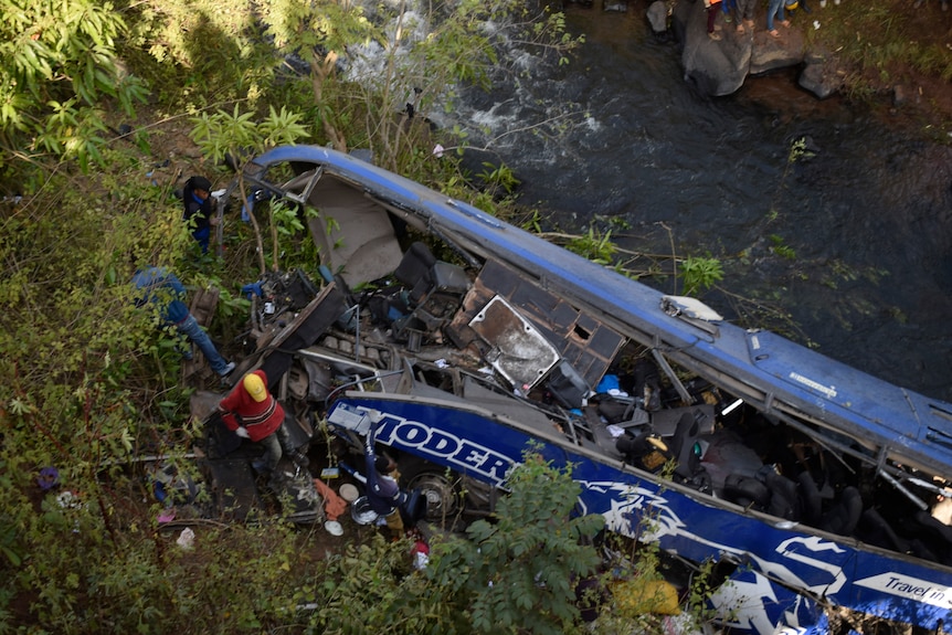 An aerial view of a blue bus crashed on the side of a river. The windows and front half are destroyed