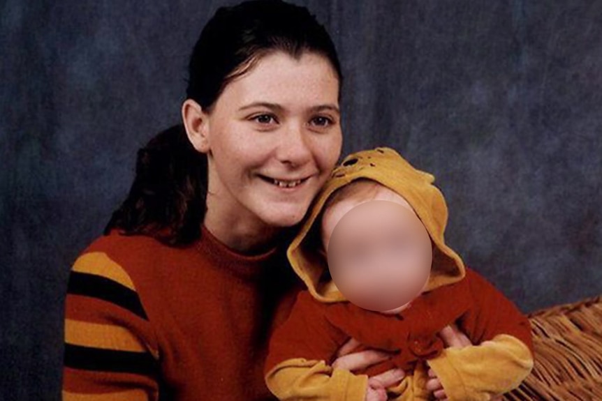 Young smiling woman with long dark hair pulled back, holding a baby, whose face is blurred. both wearing brown, mustard clothes