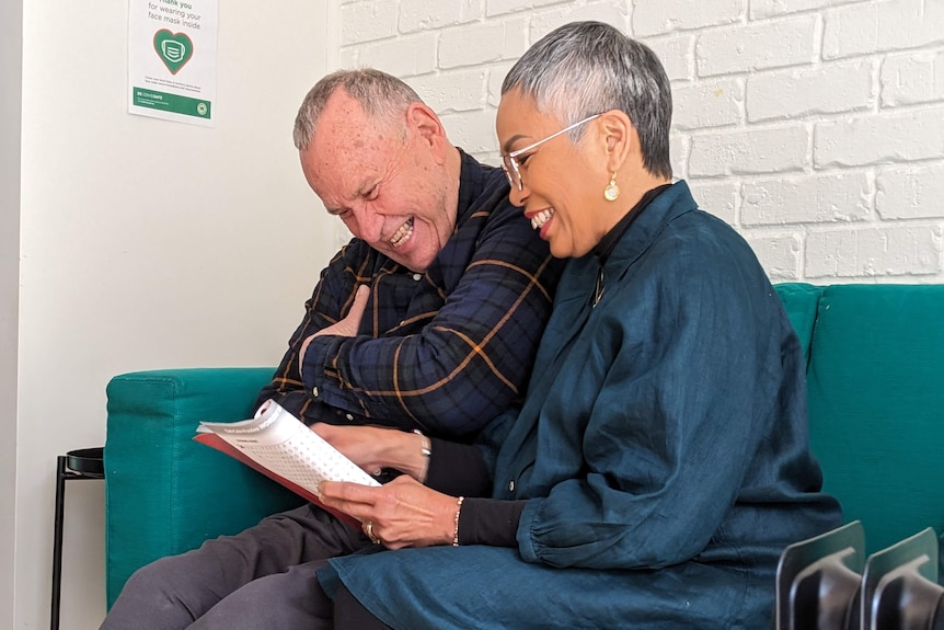 A man and woman with grey hair sit on a teal couch laughing at a book the woman is holding.