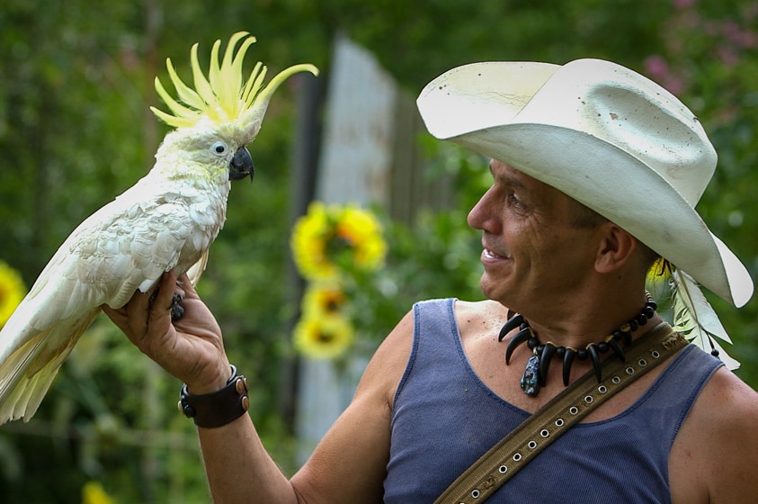 Man in a white cowboy hat is looking at a yellow-crested white cockatoo he is holding in his hand.
