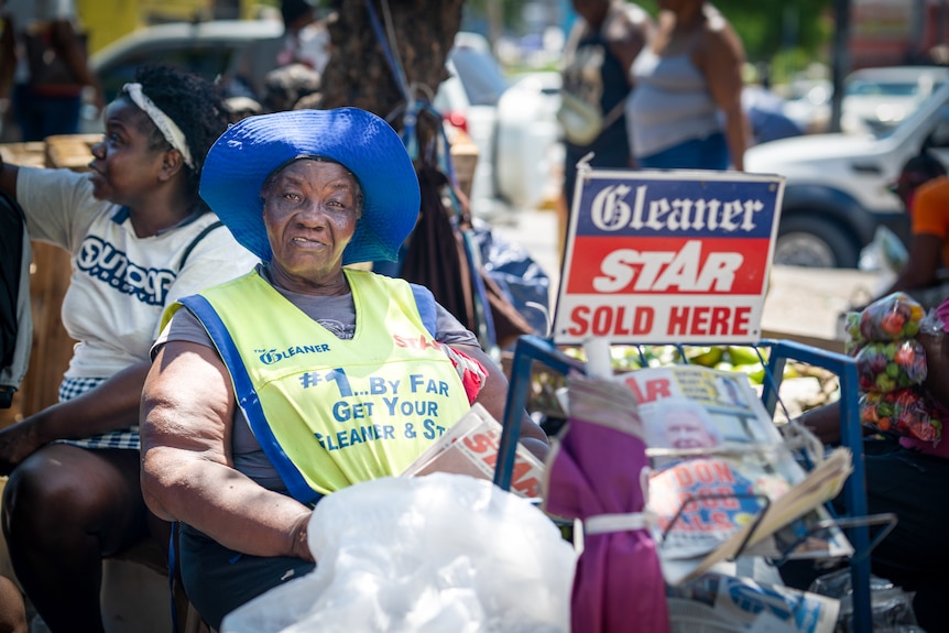 A woman in a blue hat sits next to a small newspaper stand