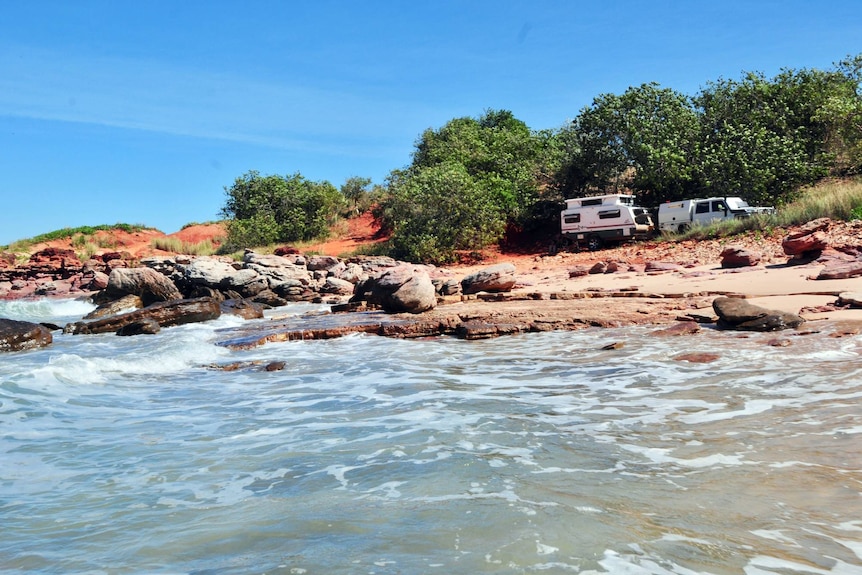 A vehicle and a camper trailer near the ocean with red dirt in the background.