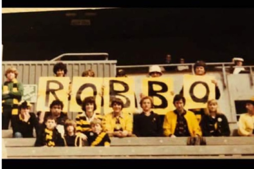 A grainy, old photograph shows a group of people gathered in the stands of a football oval dressed in yellow and black.