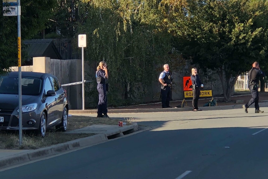 Four police officers stand on road, on phone or standing, looking at crime scene. Parked car and soft drink cans sit on curb.