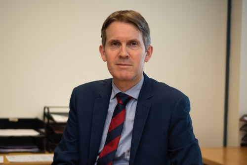 John Chesterman sits at a desk in an office wearing a suit and tie.