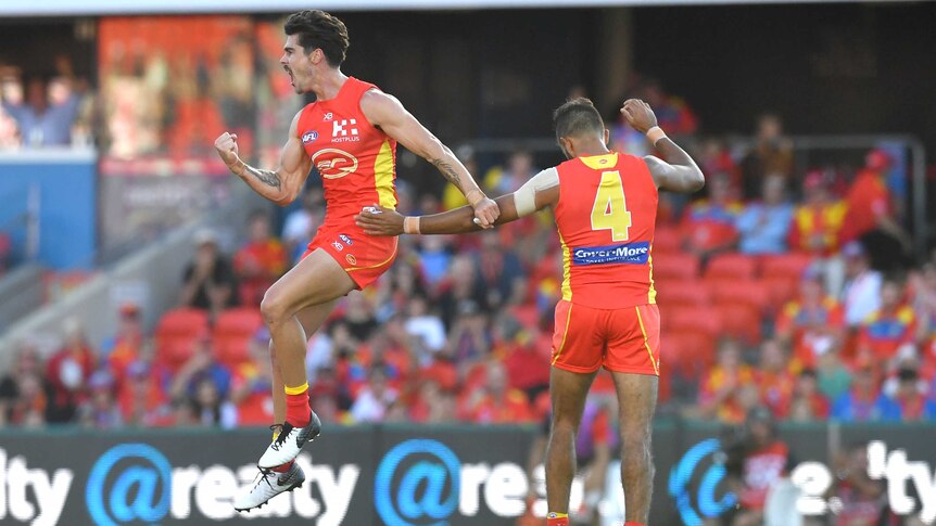 An AFL player leaps in the air in celebration after kicking a goal, as his teammate watches on.