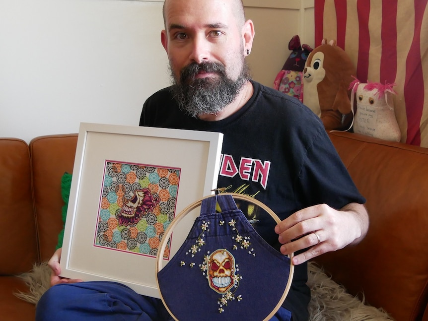 A bearded man holds up two pieces of his artwork - skulls made through embroidery
