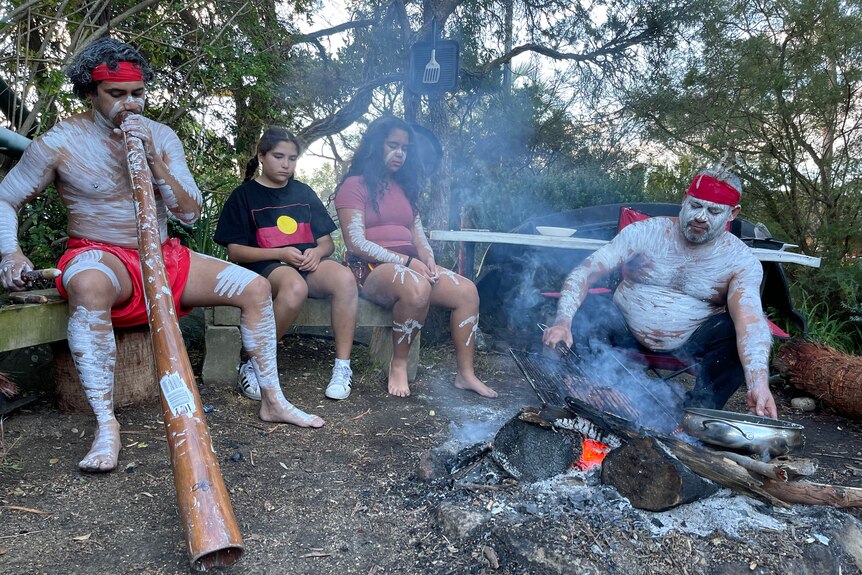 A man in traditional Aboriginal dress cooks over a fire with his two daughters watching. Another man plays a didgeridoo