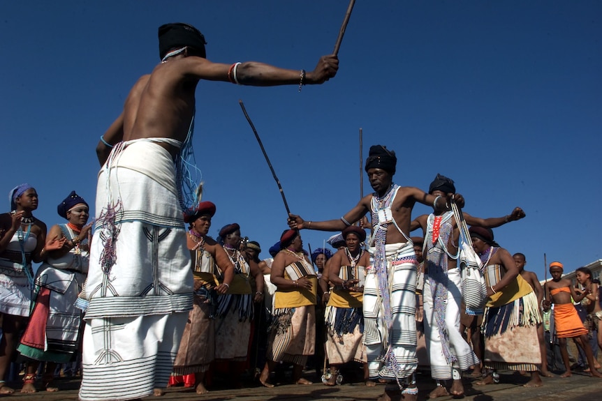 Men hold sticks or bows and wear white cloths around their chest and waists, with arms outstretched. A crowd watches.