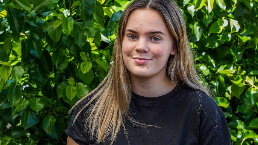 A teenage girl looks to the camera with a slight smile on her face as she stands in front of a green shrub.