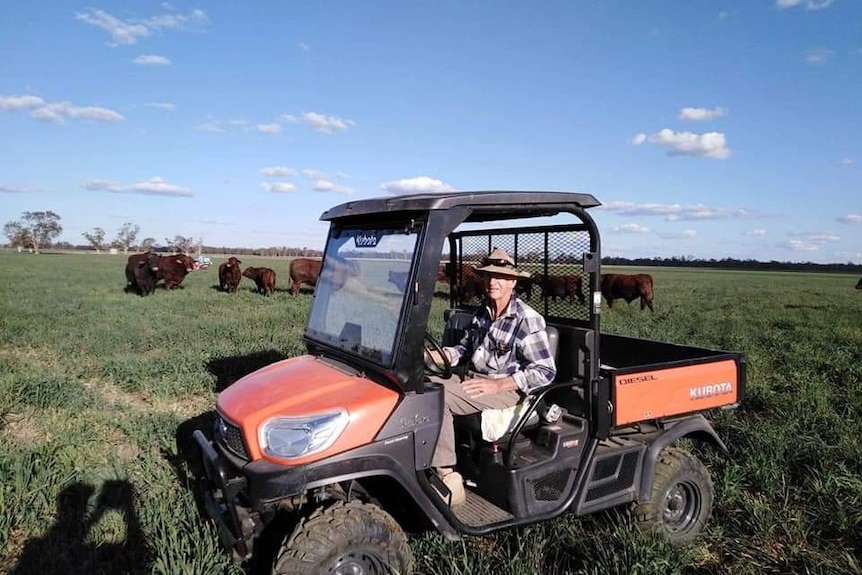 An image of Robert officer with a hat on, sitting in flannel top and brown pants in a Kubota in a paddock with cows behind him