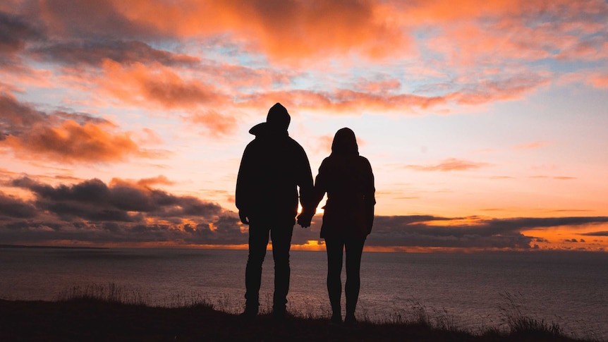 Two adults in silhouette looking at a sunset and holding hands
