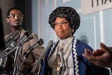 Regina looks at journalists not seen on screen as she stands in front of several mics wearing vintage clothing and glasses.