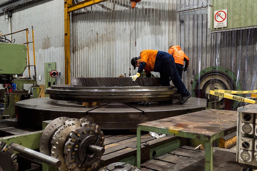 Two workers in hi-vis clothing lean over a large round machine in a workshop.