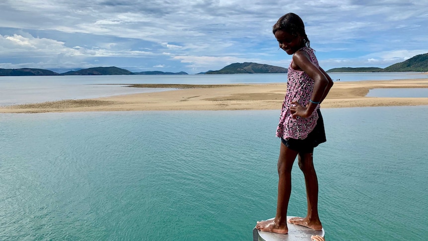 Aboriginal girl stands on podium at jetty smiling with hands on hips looking down