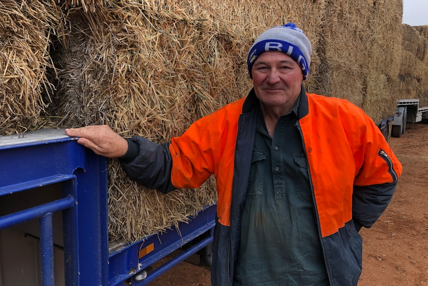 A farmer wearing a beanie and an orange hi-vis vest stands in front of bales of hay that are loaded on a low-loader