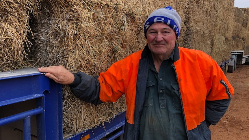 A farmer wearing a beanie and an orange hi-vis vest stands in front of bales of hay that are loaded on a low-loader