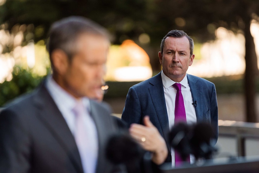 Roger Cook blurred in foreground with Premier Mark McGowan looking on.