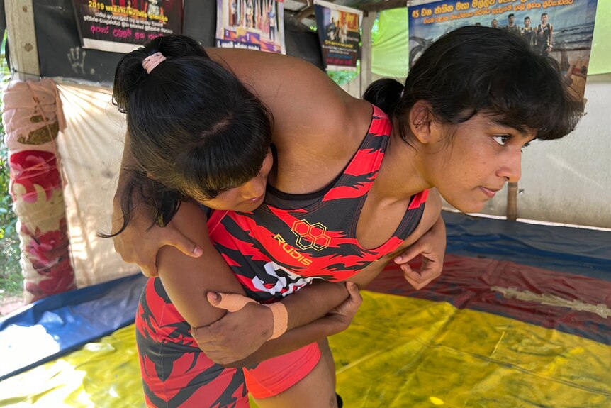 A young female wrestler wearing a red and black bodysuit had another female wrestler in a headlock.