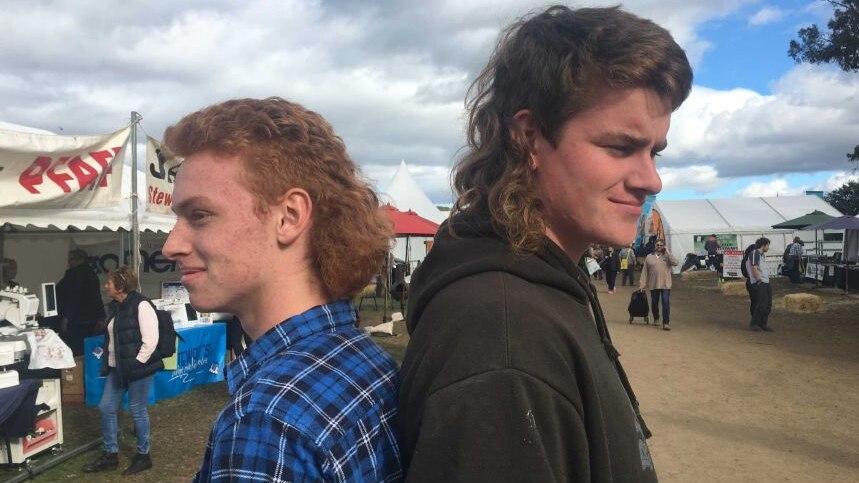 two young men standing back to back both sporting mullets one with a flannelette shirt and the other wearing a dark grey jumper