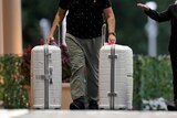 A close-up shot of a man from the waist down walking outside a hotel pulling two large white suitcases on wheels.