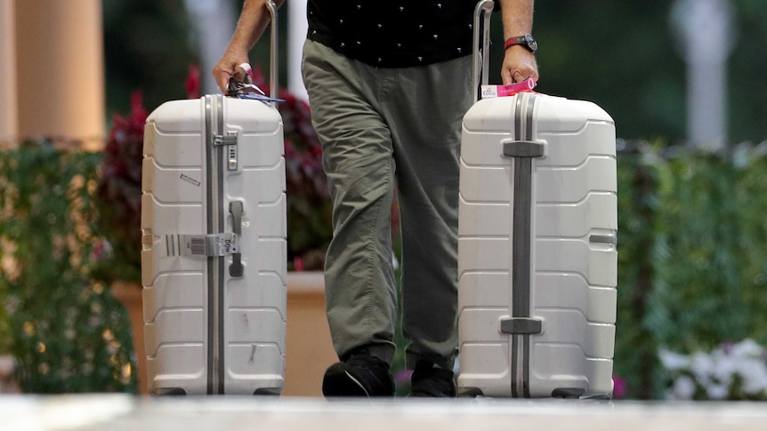 A close-up shot of a man from the waist down walking outside a hotel pulling two large white suitcases on wheels.