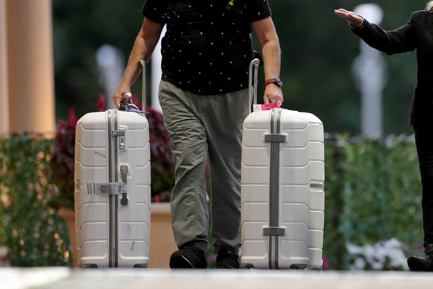 A close-up shot of a man from the waist down walking outside a hotel pulling two large white suitcases on wheels.