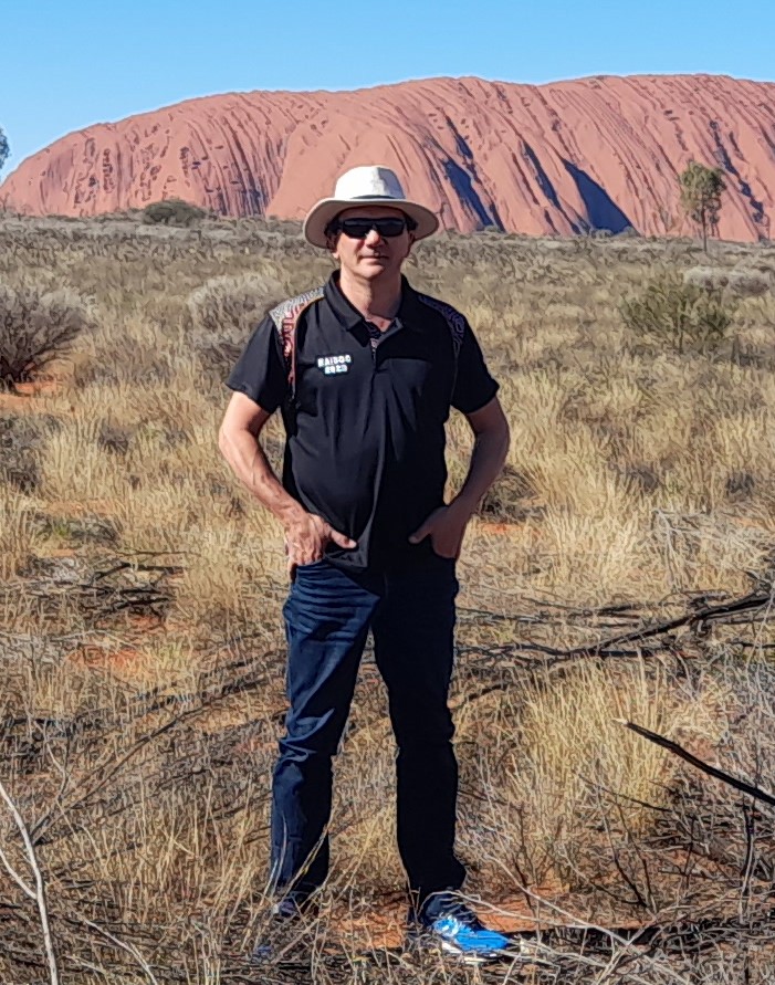 Picture of Perth Noongar man Eric Hayward in black shirt and pants in front of Uluru. 