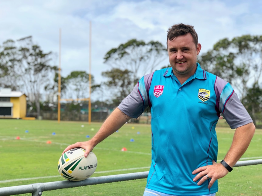 Man standing with a football, beside a football field holding a ball and smiling.