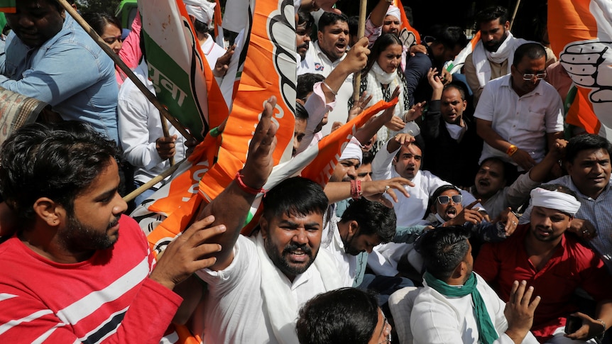 A group of Indian men and women in white traditional dress wave orange and white flags in outdoor protest