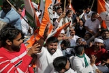 A group of Indian men and women in white traditional dress wave orange and white flags in outdoor protest