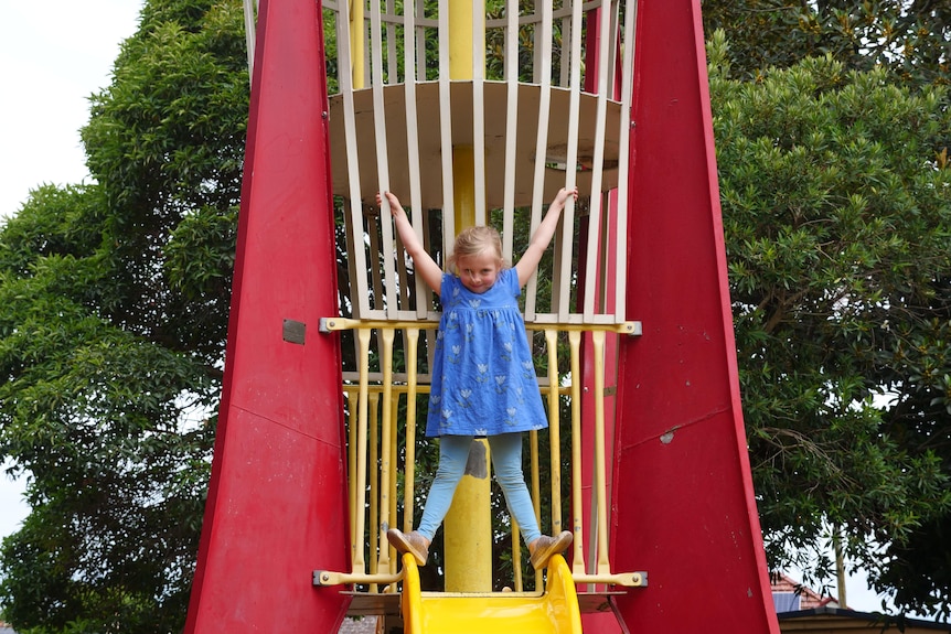 A young girl stands above a yellow slide while holding onto the bars on play equipment shaped like a rocket