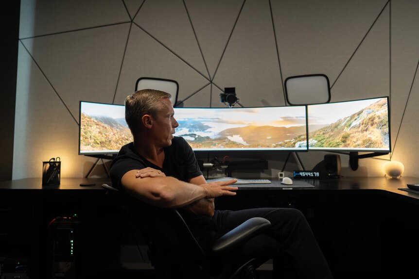 A man sits in an office chair, gesturing and looking at a massive, curved computer monitor.